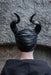 A person viewed from behind wearing a black horned headpiece or costume mask with curved horns, against a textured gray stone wall.