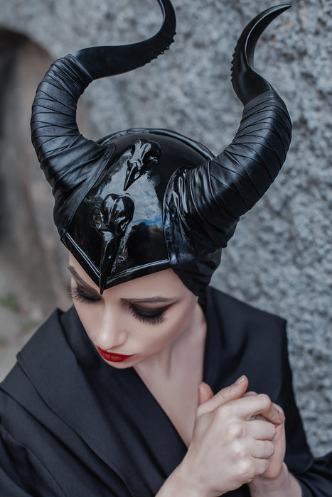 A person wearing a glossy black horned fantasy costume headpiece with curved horns, paired with a black garment, posed against a gray stone wall.