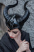A person wearing a glossy black horned fantasy costume headpiece with curved horns, paired with a black garment, posed against a gray stone wall.