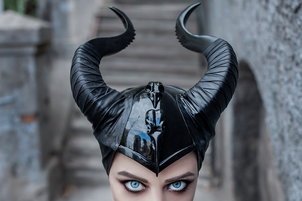 A close-up of a person wearing a glossy black horned headpiece or mask, with dramatic blue eyes and a blurred stone staircase background.