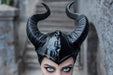 A close-up of a person wearing a glossy black horned headpiece or mask, with dramatic blue eyes and a blurred stone staircase background.