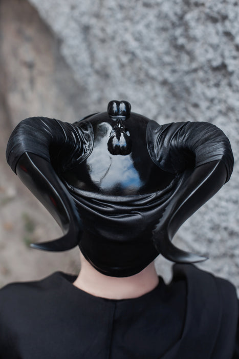 A glossy black horned mask or headpiece worn by a person, shown from the back against a textured gray stone wall background.