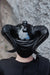 A glossy black horned mask or headpiece worn by a person, shown from the back against a textured gray stone wall background.