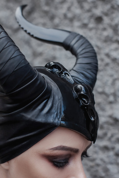 A close-up of a person wearing a glossy black horned headpiece with skull details and curved horns, against a textured gray background.