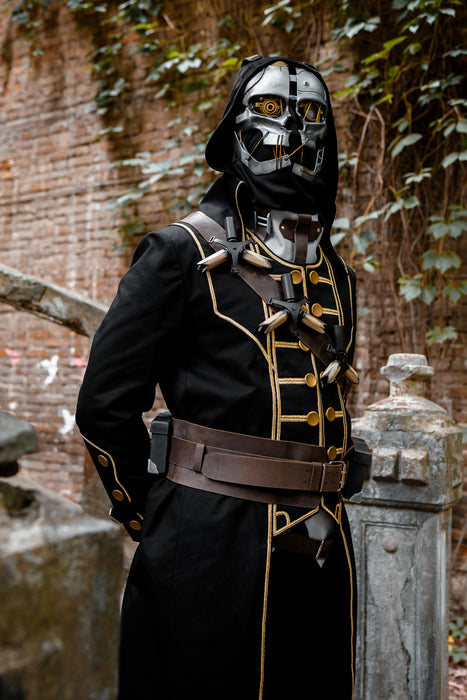 A person wearing a detailed black and silver sci-fi hooded costume with gold trim, a silver helmet with glowing orange eyes, and a brown belt, standing outdoors in front of a brick wall and stone pillar.