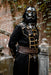 A person wearing a detailed black and silver sci-fi hooded costume with gold trim, a silver helmet with glowing orange eyes, and a brown belt, standing outdoors in front of a brick wall and stone pillar.