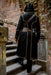 A person viewed from behind wearing a long black hooded robe with gold trim and brown belt details, walking up outdoor stone stairs against a brick wall and vines.